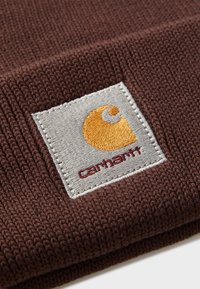 Detailní záběr na hnědou pletenou l�átku s přišitou čtvercovou nášivkou zobrazující oranžové logo a název Carhartt na béžovém pozadí.