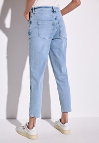 Jeans en denim bleu clair avec une jambe fuselée, taille haute et deux poches arrière. Portés avec des baskets blanches. Texture lisse, design épuré.
