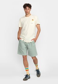 Crème t-shirt met een zak en een kleurrijk ontwerp, in combinatie met lichtgroene corduroy shorts. Sokken met meerkleurige strepen en sneakers.
