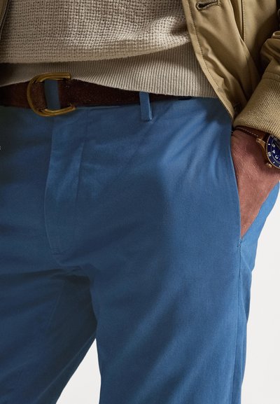 Calças azul claro com textura lisa, com bolsos laterais, cinto castanho e camisola de malha cor creme por cima.