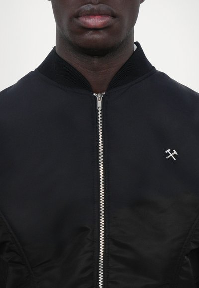 Veste en bomber noire avec fermeture éclair, col côtelé et accent logo métallique. Texture lisse avec une silhouette ajustée.