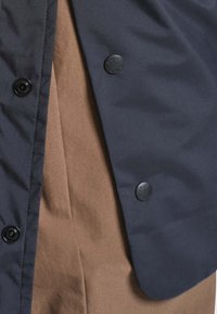 Gros plan d'une veste bleu marine avec des boutons-pression noirs sur un tissu marron texturé, montrant les détails des boutons et le contraste du tissu.