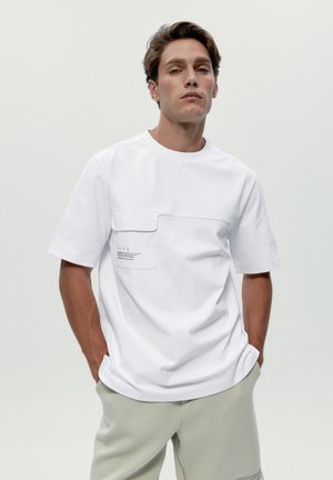 Mann trägt ein weißes Kurzarmhemd mit Brusttasche und hellgraue Hose, steht mit den Händen in den Taschen vor einem schlichten Hintergrund.