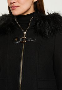 Cappotto nero con colletto in pelliccia sintetica, rifinito con zip dorata e chiusura con cinturino in pelle. Tessuto testurizzato con silhouette strutturata.