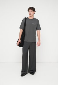 Γκρι βαμβακερό t-shirt με κοντά μανίκια και λογότυπο, σε συνδυασμό με γκρι παντελόνι με φαρδιά πόδια. Το μοντέλο κρατάει μια υφασμάτινη μαύρη τσάντα.