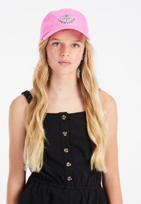 Cappellino rosa con testo e design ricamati in verde. Indossato sopra una canotta nera, con bottoni, senza maniche e con una vita elasticizzata. Capelli lunghi e ondulati visibili.