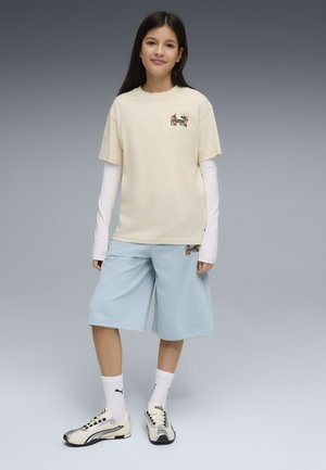 Dziewczyna z długimi ciemnymi włosami w kremowej koszulce Puma na białych długich rękawach, jasnoniebieskich szortach, białych skarpetkach Puma i kremowych sneakersach.