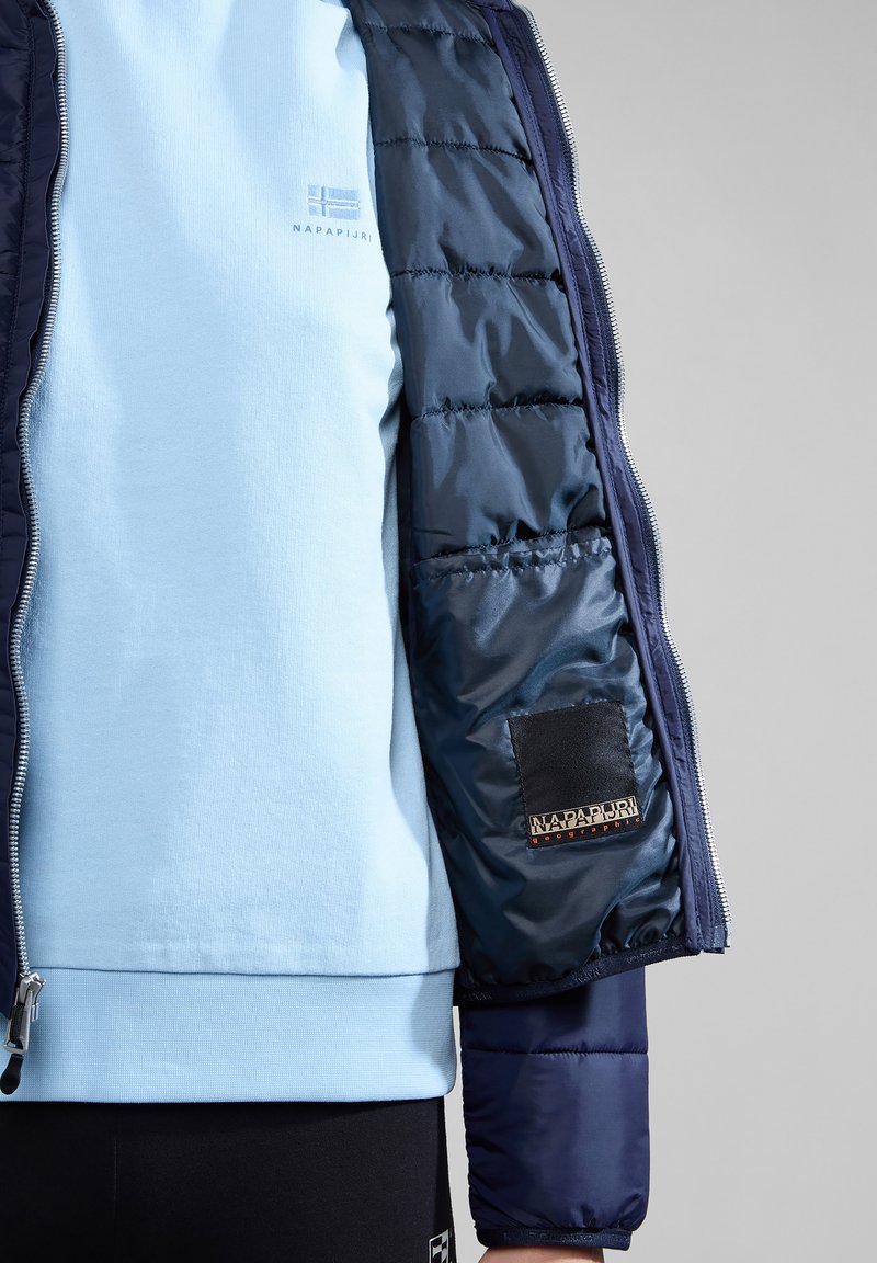 Giubbotto imbottito di colore blu navy con finitura trapuntata, abbinato a una maglietta azzurra con collo a giro. Presenta un'etichetta di marca e dettagli con cerniera.