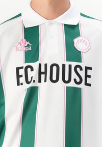 Valkoinen poolopaita, jossa on vihreitä ja vaaleanpunaisia pystysuoria raitoja. Vasemmalla puolella on musta "F.C. HOUSE" -logo ja vaaleanpunainen Kappa-embleemi.
