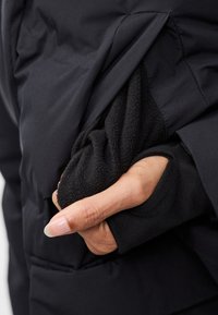 Mano parzialmente inserita nella tasca di una giacca nera foderata in pile, che mostra maniche sovrapposte con foro per il pollice sul capo interno nero.