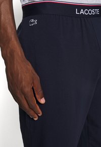 Námornícke modré bavlnené tepláky s elastickým pásom, na ktorých je biele logo Lacoste, hladká textura a priliehavý strih.