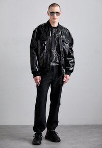 Casaco bomber preto brilhante com dois bolsos no peito, combinado com um sweatshirt gráfico preto e calças cargo a condizer. Ténis pretos completam o visual.