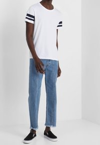 Mand iført hvid T-shirt med sorte ærme-striber, slidte blå jeans og sorte slip-on sneakers, stående mod en hvid baggrund.