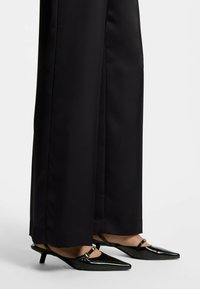 Black slingback heels with a pointed toe, shiny patent finish, and small heel, paired with wide-leg black trousers.