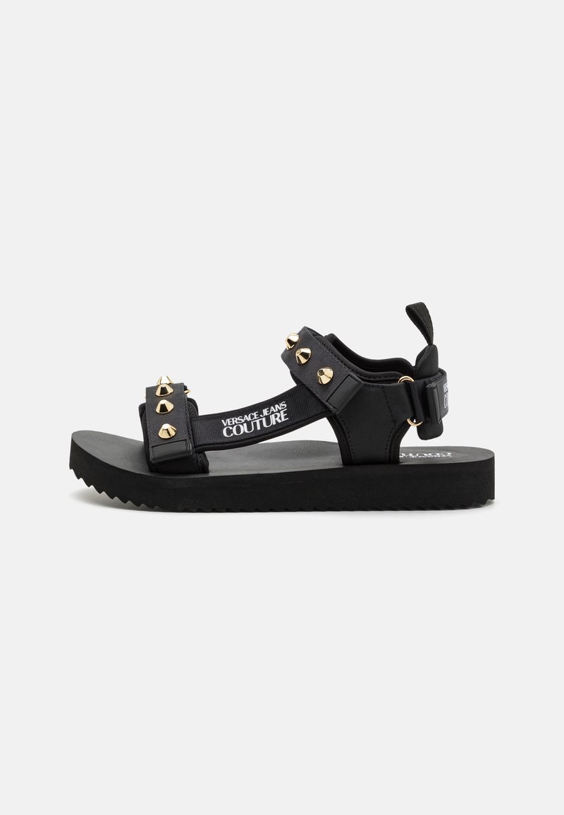 Svarte flate sandaler med brede stropper, utstyrt med gullnitter og en teksturert såle. Merkevaren sin logo er synlig på stroppen. Uformelt design.