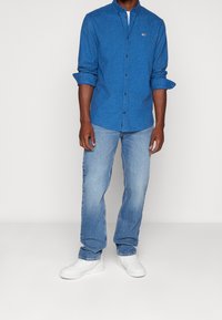 Blaues, strukturiertes Hemd mit Knopfleiste und kleinem Logo, kombiniert mit hellblauen Jeans und weißen Sneakers, vor einem einfarbigen Hintergrund stehend.