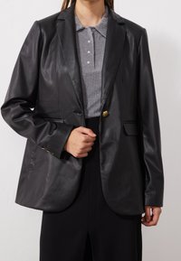 Blazer en cuir noir avec col cranté, fermeture à un bouton, et un haut en maille grise texturée en dessous. Présente des accents en métal doré.