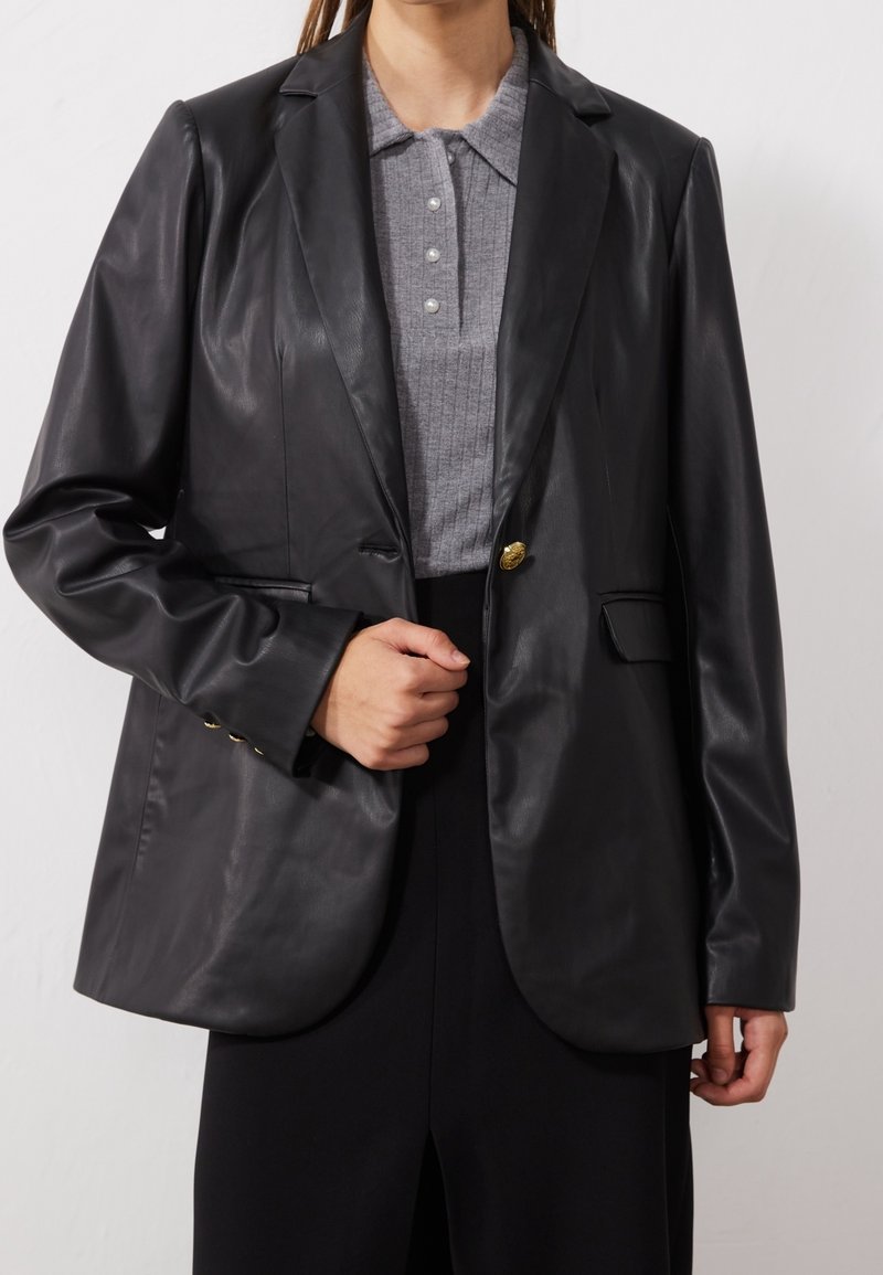 Blazer en cuir noir avec col cranté, fermeture à un bouton, et un haut en maille grise texturée en dessous. Présente des accents en métal doré.