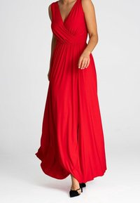 Femme portant une robe maxi rouge sans manches avec un décolleté en V profond et des chaussures à talons hauts noires marchant vers l'avant sur un fond uni.