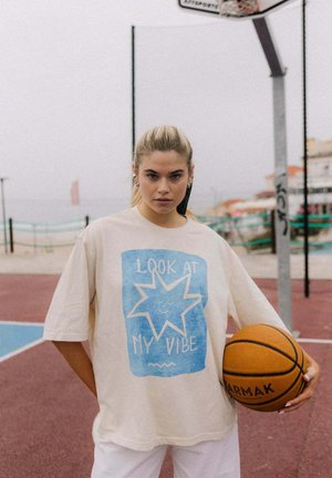 Young woman holding basketball stands on outdoor court wearing beige shirt with blue star and text "Look at my vibe."