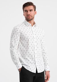 Camisa blanca de botones con patrones impresos en negro, mangas largas, cuello y corte recto. Textura suave con un diseño clásico.