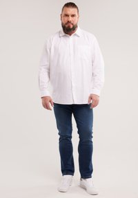 Man with beard and tied-back hair wearing a white button-up shirt, blue jeans, and white sneakers standing against a plain background.