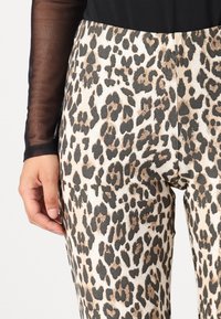 Oroszlánmintás leggings sima, textúrált felszínen; világosbarna alap fekete és barna foltokkal; áttetsző fekete, hosszú ujjú toppal párosítva.