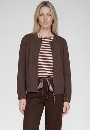 Blonde Frau trägt eine braune Jacke mit Reißverschluss über einem rosa-braun gestreiften Hemd, dazu braune Hosen, die mit einem rosa Schal in der Taille gebunden sind.