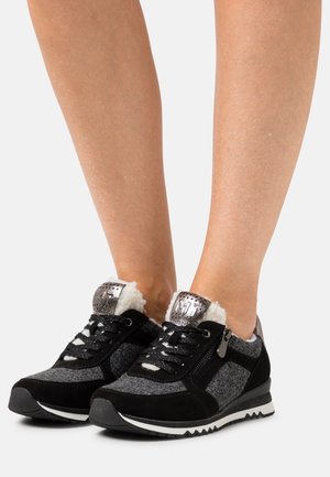 Füße, die schwarze und graue Schnürsneakers mit weißem Fleecefutter und metallischen Akzenten tragen, auf weißem Hintergrund stehend.