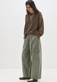 Brun strikket sweater over løse, olivengrønne cargos med sidelommer; modellen bærer mørke sko med en lys accent. Enkel baggrund.