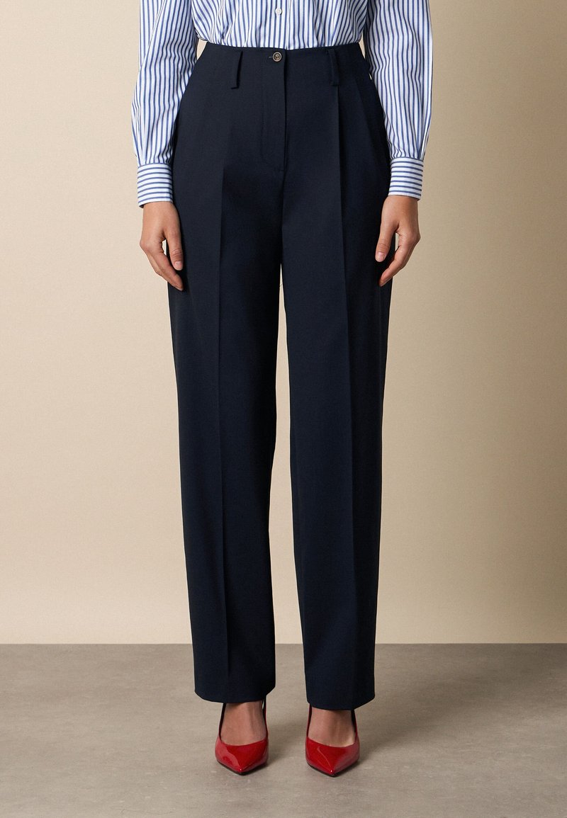 Vrouw die marineblauwe, op maat gemaakte broek met hoge taille, blauw-wit gestreept overhemd met knopen en rode puntenhakken draagt, staand op een grijze vloer.