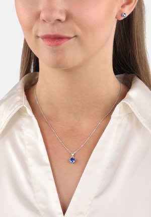 Collana d'argento con un pendente di gemma blu, indossata con una camicia bianca con colletto. Piccole orecchini a dado blu completano il look.