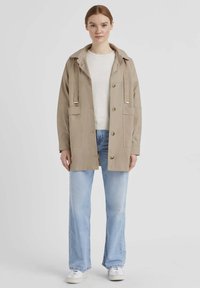 Beige leichte Jacke mit Kapuze, vorderen Knöpfen, zwei großen Taschen und verstellbaren Riemen. Kombiniert mit einem weißen Pullover und hellblauen Jeans.