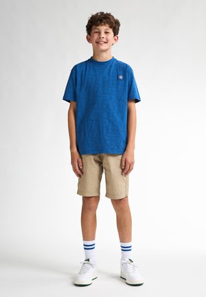 Blaue kurzärmlige T-Shirt mit strukturiertem Schriftzug, khakifarbene Shorts, weiße Sneaker mit grünen Akzenten und gestreifte weiße Socken.