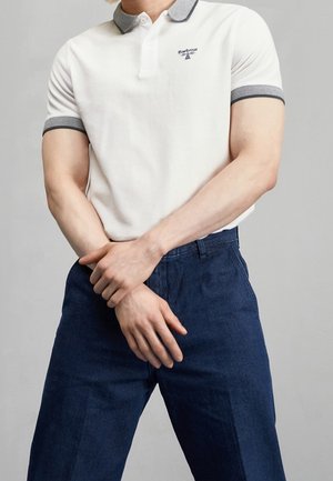 Mann trägt weißes kurzärmeliges Poloshirt mit grauem Kragen und Manschetten, dunkelblaue Hose, steht mit verschränkten Händen vor dem Körper.
