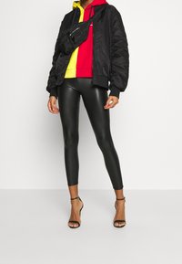 Giacca bomber nera sopra una felpa con cappuccio rossa e gialla, abbinata a leggings neri lucidi e sandali neri con cinturini. Marsupio indossato a tracolla.