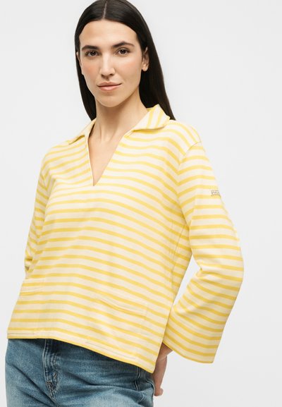 Γυναίκα με μακριά σκούρα μαλλιά που φοράει μία κίτρινη και λευκή ριγέ μπλούζα με μακριά μανίκια και μπλε τζιν, στέκεται μπροστά σε ένα απλό φόντο.