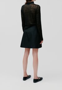 Schwarzes langärmeliges Oberteil mit transparentem Stoff, kombiniert mit einem schwarzen A-förmigen Rock. Modell trägt flache schwarze Schuhe, Ansicht von hinten.