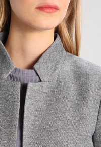 Chaqueta de mezcla de lana gris con un cuello alto y tejido texturizado, llevada encima de una blusa de tono púrpura claro con canalé. Vista en primer plano del escote.