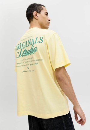 Mand iført en løs, blekgul T-shirt med stor turkis tekst "Originals Studio" på ryggen, kombineret med mørke bukser mod en hvid baggrund.