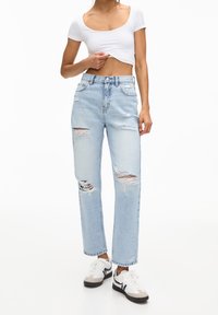 Jeans de mezclilla decolorados y rasgados, con pierna recta, cintura alta y múltiples áreas desgastadas, combinados con zapatillas blancas.