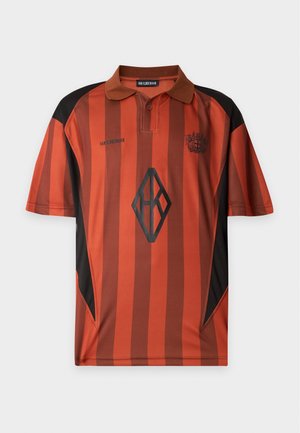 Un homme se tient les mains dans les poches, portant une chemise à manches courtes rayée orange et noire et un pantalon cargo marron ample avec des chaussures noires.