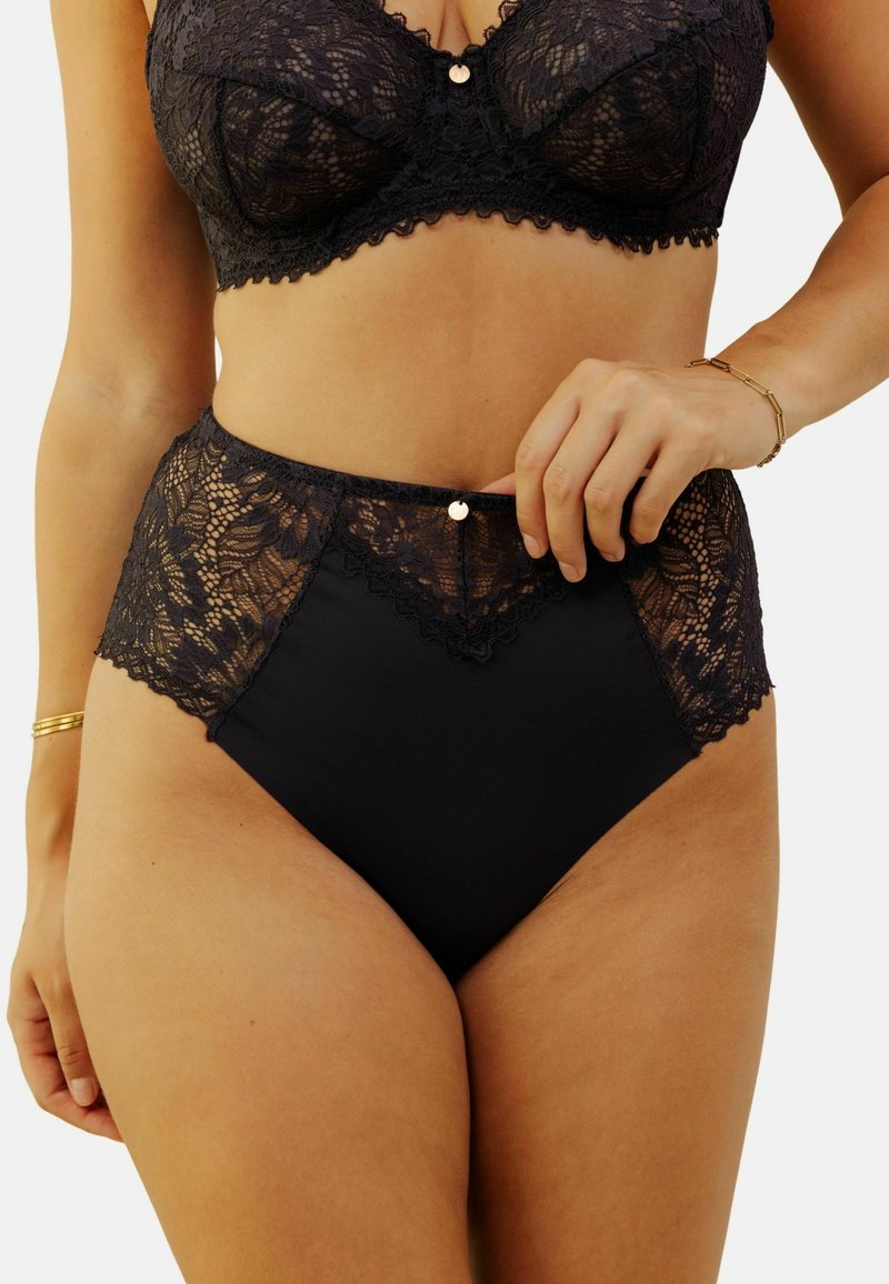 Lingerie noire taille haute avec des accents en dentelle et un bord festonné, présentant un petit bouton métallique au centre devant et un tissu lisse.