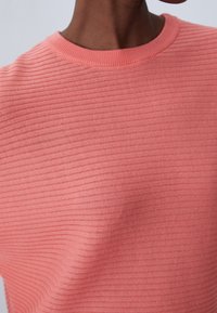 Ribbed, dzianinowy top w miękkim koralowym kolorze, z okrągłym dekoltem i fakturowanym materiałem z poziomymi paskami. Bez widocznych elementów metalowych ani akcentów.