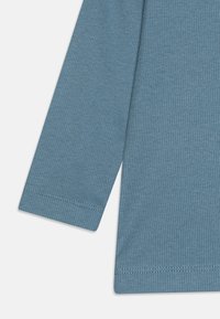 Langermet ribbet topp i mykt blått materiale. Har innfelte sømmer og en glatt tekstur, med en tettsittende design og smale ermer.