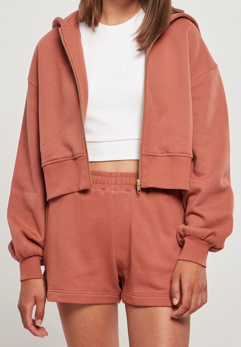 Felpa corta color rosa con zip, ampie tasche e polsini a coste, abbinata a pantaloncini coordinati. Realizzata con tessuto morbido e confortevole.