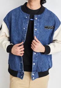 Kurtka bomberka z denimu w kolorze niebieskim z kremowymi rękawami, prążkowanymi mankietami i zatrzaskami. Na piersi znajduje się czarne logo "HUGO".