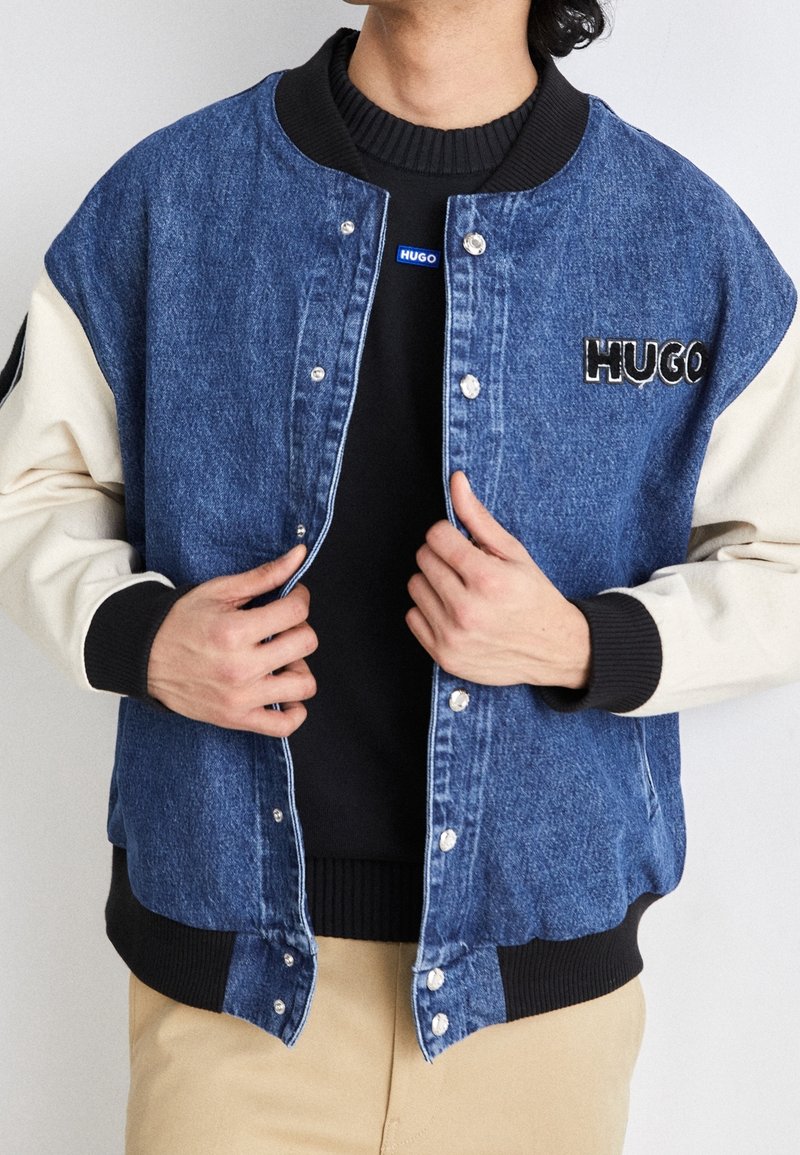 Kurtka bomberka z denimu w kolorze niebieskim z kremowymi rękawami, prążkowanymi mankietami i zatrzaskami. Na piersi znajduje się czarne logo "HUGO".