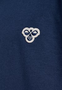 Logo d’abeille brodé en blanc et bleu marine sur fond de tissu bleu foncé texturé.