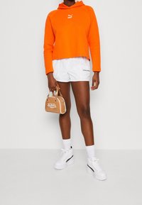 Persoon in een heldere oranje Puma hoodie en witte Puma shorts, met een beige Von Dutch handtas, staande met witte sneakers en sokken.